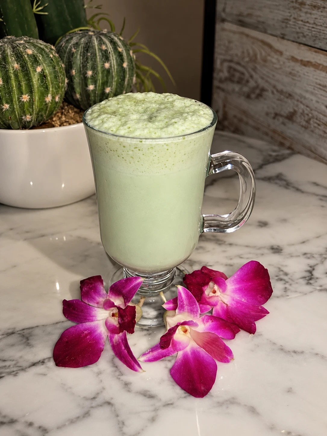 Frothy green drink in a glass mug beside purple orchids and cactus plants